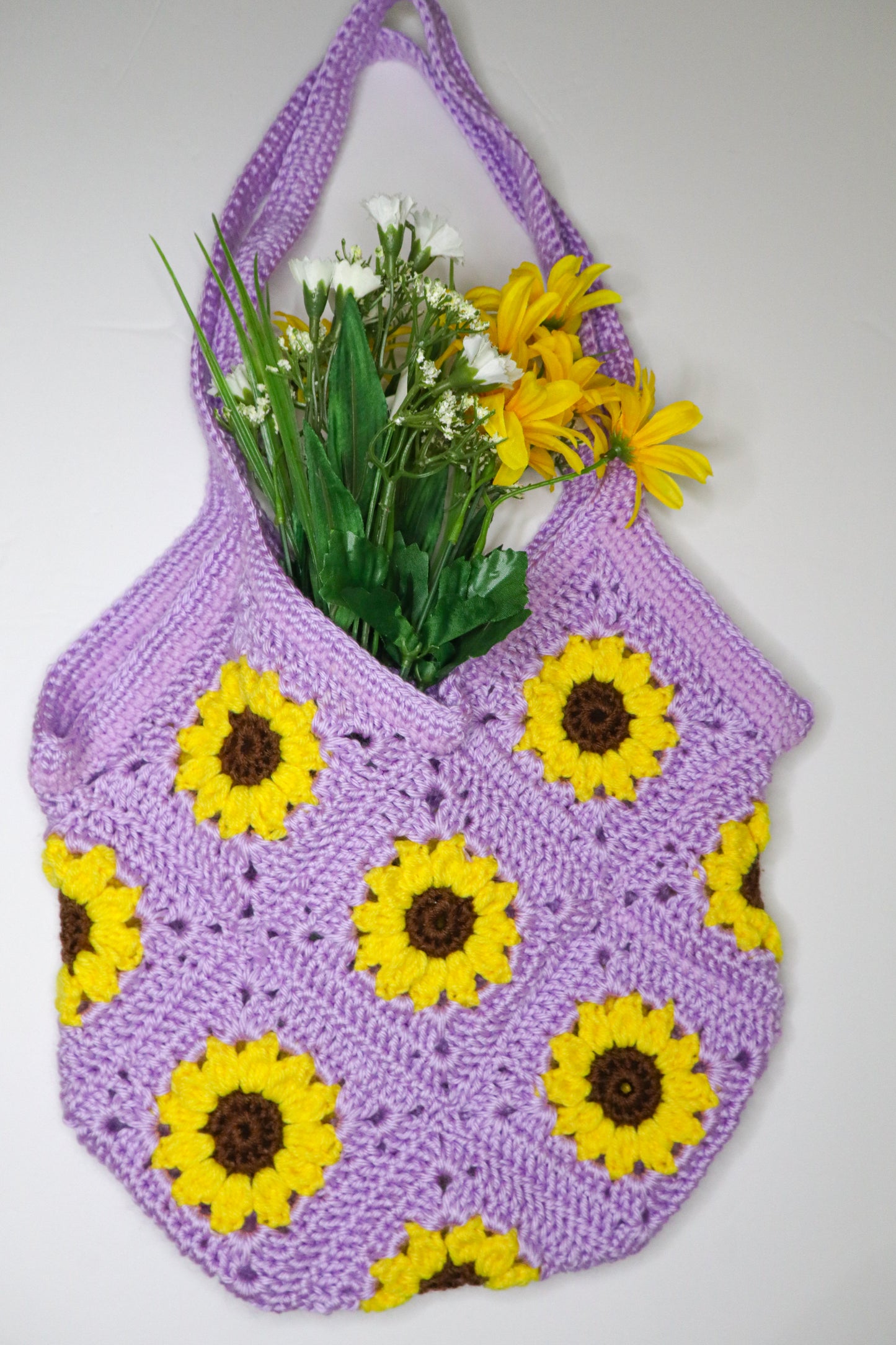 Purple crochet bag with yellow sunflower patterns filled with flowers on a white background